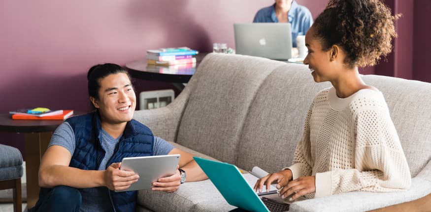 two people sitting around a sofa reading on a computer and tablet